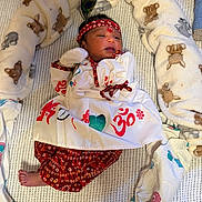 Prisha is registered to the contest to win money with this photo: newborn, baby, infant, blanket, headband, peacock_feather, red_clothing, white_clothing, patterned_clothing, cushion, plush_toy, animal_print, bed, lying_down, indoors, portrait, cute, soft_texture, decorative, peaceful