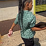 child, braided_hair, green_shirt, black_pants, tablet, sidewalk, sunlight, urban, yellow_door, metal_shutter, footwear, walking, outdoor, shadow, person, casual_clothing, handheld_item, concrete, street, daylight