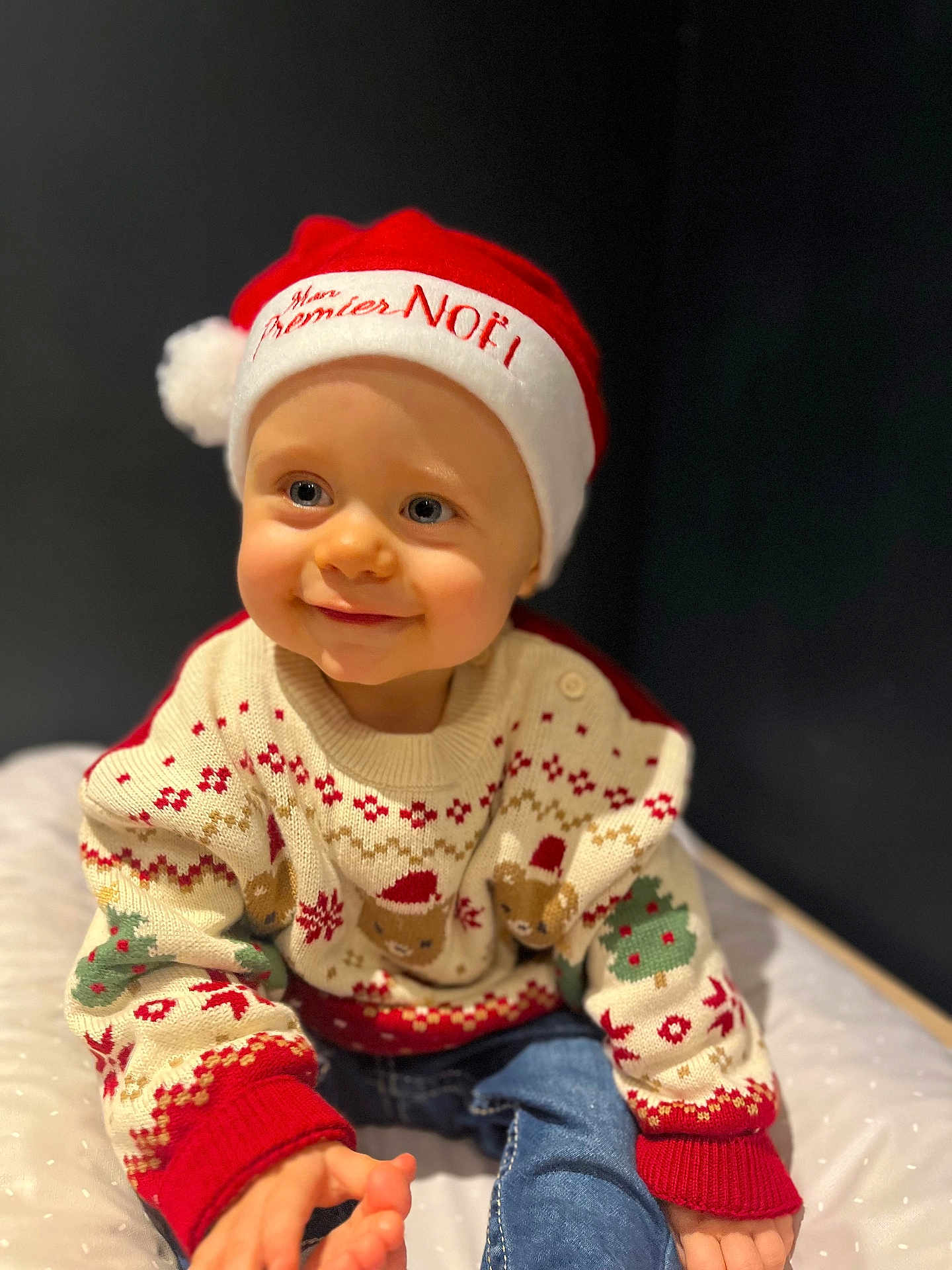 Sacha participe au concours pour gagner de l'argent avec cette photo : baby, child, smiling, santa_hat, christmas_sweater, festive, holiday, cute, portrait, indoor, clothing, jeans, happy, person, infant, face, headwear, seated, soft_surface, dark_background