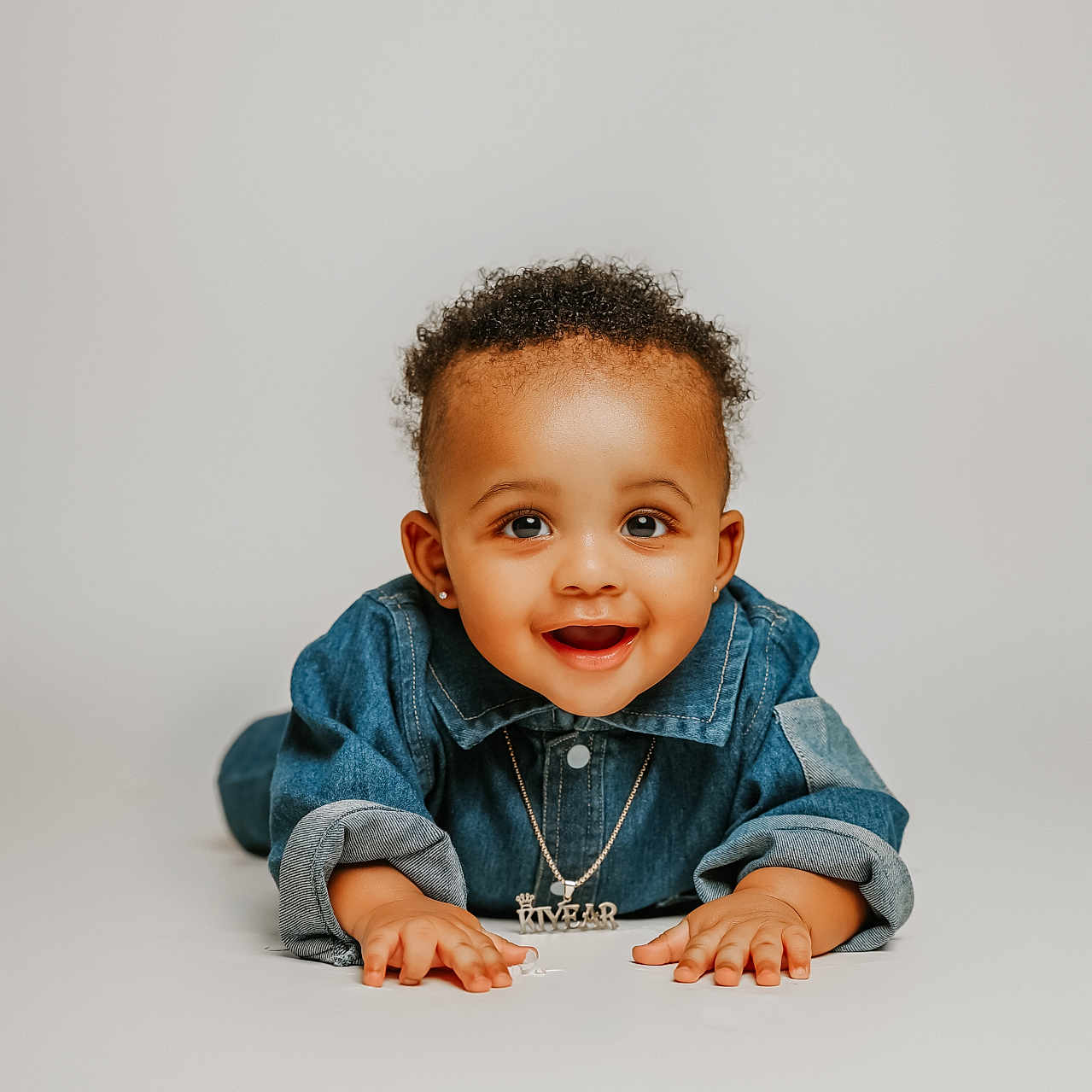 Kiyear is registered to the contest to win money with this photo: baby, child, smiling, denim_jacket, curly_hair, earrings, portrait, studio, neutral_background, happy, cute, infant, lying_down, hands, face, young, fashion, jewelry, person, expression