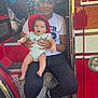 baby, child, woman, fire_truck, helmet, red, outdoor, person, holding, sitting, footwear, shadow, step, clothing, smile, expression, vehicle, metal, casual, daylight
