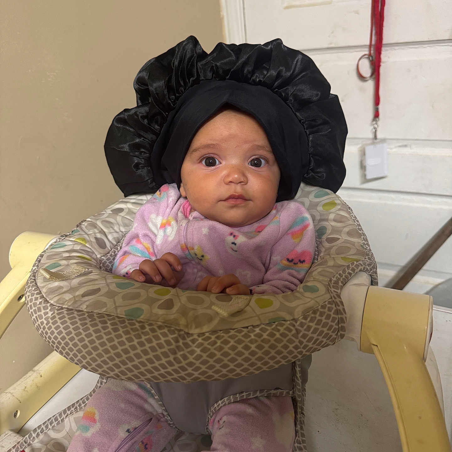 Soraya joined the competition — help win amazing prizes! baby, baby_seat, black_bonnet, child, curious, cute, door, face, furniture, hands, headwear, home, indoors, infant, lanyard, pajamas, patterned_fabric, pink, seated, wide_eyes