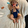baby, infant, child, portrait, lying_down, blanket, crocheted_hat, overalls, knitwear, toy, red_toy, white_blanket, indoors, close_up, face, hands, feet, smiling, soft_lighting, cozy