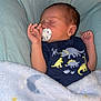 baby, newborn, sleeping, pacifier, onesie, dinosaur_print, blanket, cushion, chair, infant, peaceful, cozy, soft, cute, child, indoors, resting, hand, face, portrait