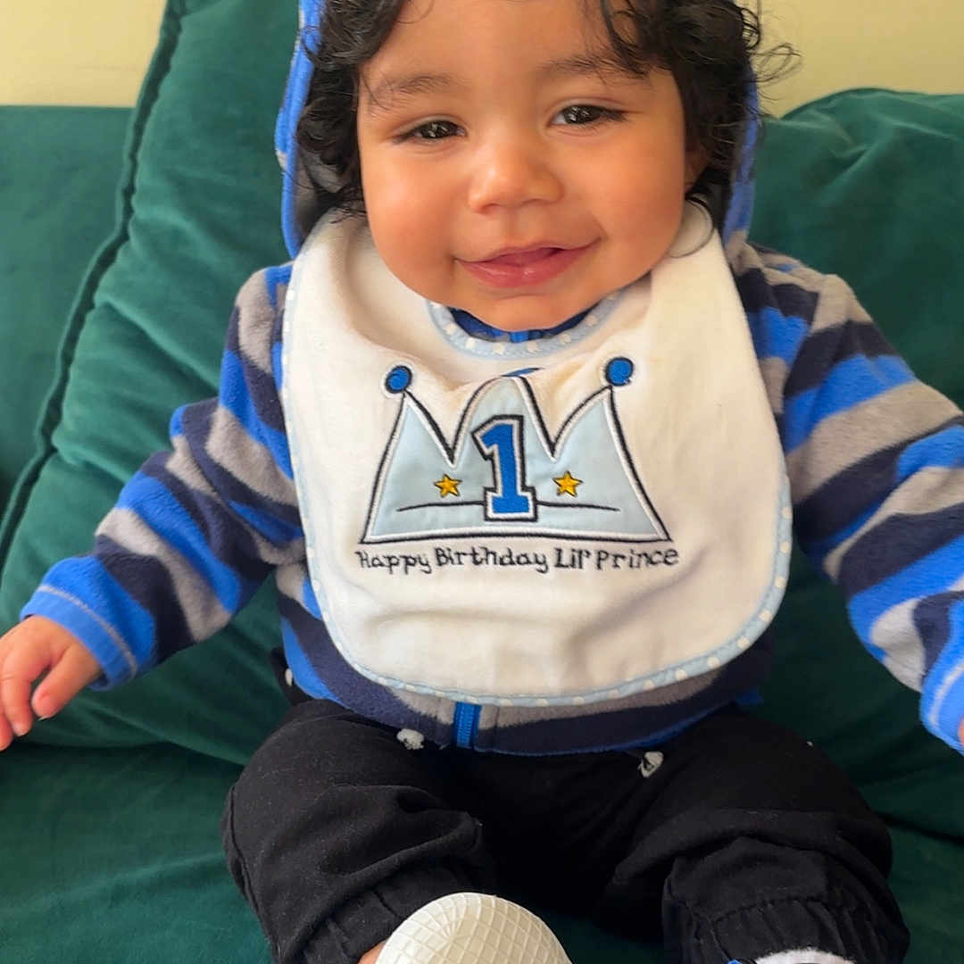 Tairell is registered to the contest to win money with this photo: baby, bib, birthday, black_pants, child, clothing, couch, curly_hair, cute, face, footwear, green_couch, happy, hoodie, indoor, portrait, sitting, smiling, white_shoes, young