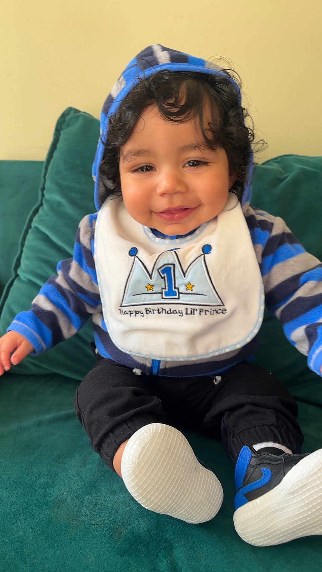 Tairell is registered to the contest to win money with this photo: baby, bib, birthday, black_pants, child, clothing, couch, curly_hair, cute, face, footwear, green_couch, happy, hoodie, indoor, portrait, sitting, smiling, white_shoes, young