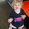Laura is registered to the contest to win money with this photo: toddler, child, smiling, curly_hair, blonde, blue_eyes, black_tshirt, pink_shorts, indoor, playground, carpet, happy, person, footwear, cute, young_child, reaching_out, play_area, colorful