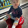 baby, banner, casual, child, colorful_clothing, curly_hair, cute, floor_carpet, heart_pattern, indoor, mesh_screen, navy_blue_top, person, playful, portrait, red_stool, sitting, smiling, toddler, young_child