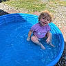 toddler, child, pool, water, outdoor, sunny, blue, grass, gravel, curly_hair, purple_shirt, splashing, happy, smiling, summer, play, plastic_pool, casual_clothing, nature, daylight