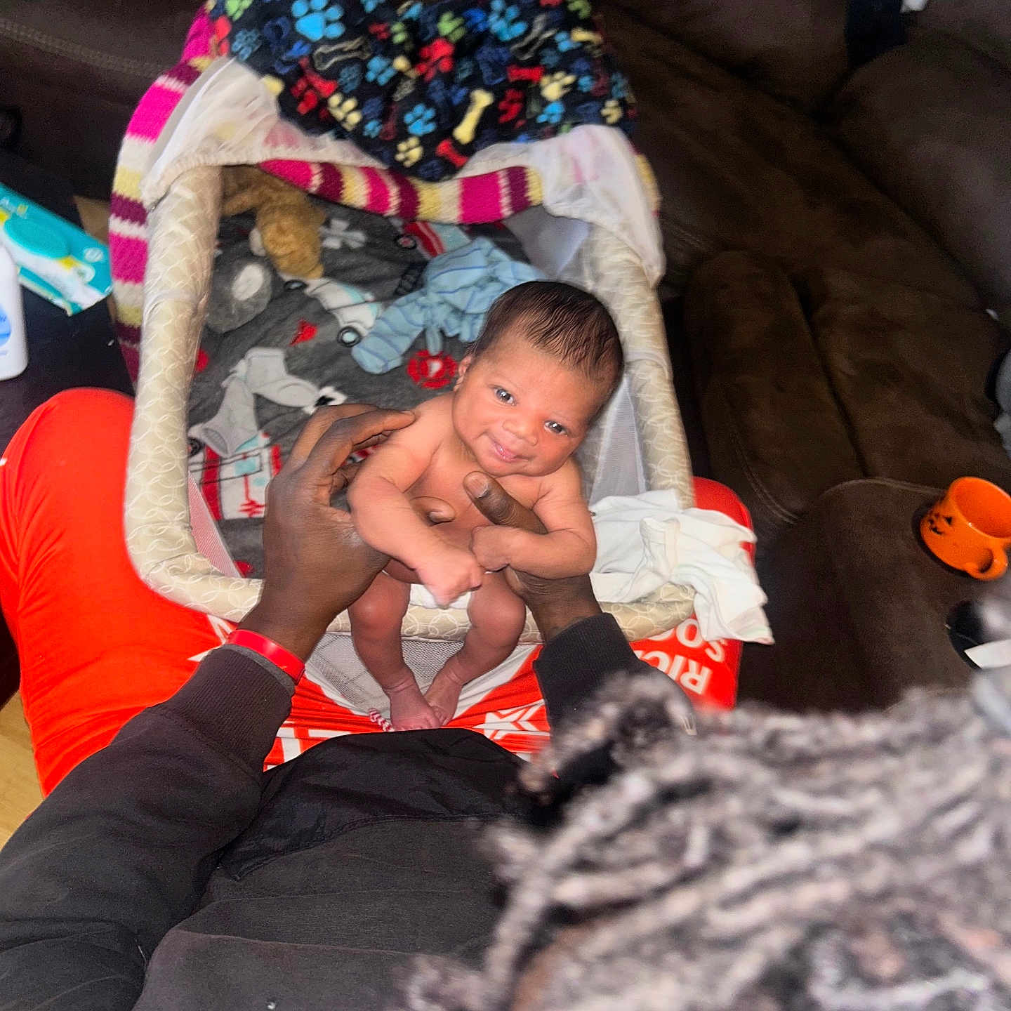 Errol is registered to the contest to win money with this photo: adult, baby, baby_bassinet, black_jacket, blanket, clothing, couch, furniture, hair, hands, head, holding, indoor, infant, orange_clothing, person, room, sitting, skin, smile