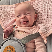 Lucie a rejoint le concours — aidez-le/la à gagner de superbes lots ! baby, infant, smiling, drooling, pink_blanket, baby_bouncer, safety_harness, onesie, face, hand, leg, ear, soft_texture, plush, cozy, portrait, home_interior, radiator, brand_label, gray_fabric
