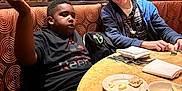 Nicholas is registered to the contest to win money with this photo: child, boy, restaurant, table, plate, food, utensils, booth, mirror, reflection, casual_clothing, jacket, tshirt, indoor, seated, expression, gesture, brown_hair, black_hair, friendship