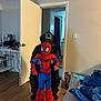 child, costume, spiderman, indoor, person, doorway, floor, blanket, bag, wood_floor, clothing, black_hood, toy, casual, home, curtain, door, socks, play, holding