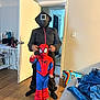 child, costume, spiderman, adult, black_clothing, hood, indoor, door, floor, wooden_floor, bag, blanket, furniture, person, pose, curtain, room, socks, toy_shoes, light_switch