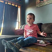 Lanie is registered to the contest to win money with this photo: toddler, child, indoor, living_room, table, boots, rain_boots, pink_shirt, curtains, sofa, carpet, wooden_floor, window, natural_light, portrait, expression, decor, photograph, toy, rock