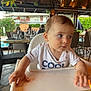 baby, child, high_chair, restaurant, table, indoor, people, furniture, glass, plant, outdoor, dining, blur, hand, shirt, text, face, seat, wood, casual