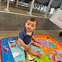 Elias Mikhaël participe au concours pour gagner de l'argent avec cette photo : baby, child, play_mat, colorful, indoor, floor, shirt, shorts, person, legs, chairs, shopping_bag, curious, seated, expression, footwear, background, animal_print, toddler, candid