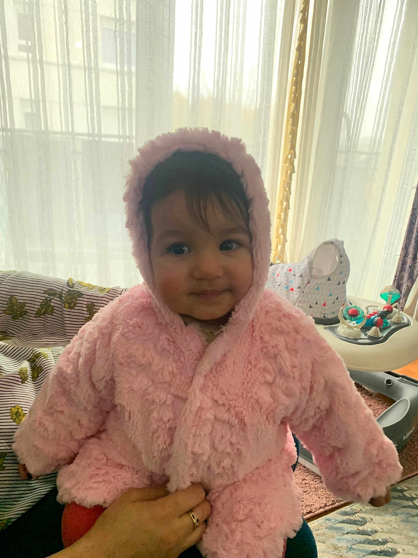 Jana participe au concours pour gagner de l'argent avec cette photo : baby, child, pink_jacket, hooded_clothing, indoor, curtains, hand, person, supporting_hand, face, eyes, smile, furniture, carpet, baby_walker, toy, window, light, cozy, portrait