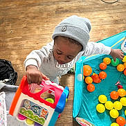Dior is registered to the contest to win money with this photo: child, toy, beanie, sweatshirt, playpen, plastic_balls, floor, wooden_floor, indoor, playtime, colorful, person, young_child, activity, hand, face, headwear, casual_clothing, room, play