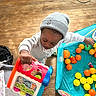 child, toy, beanie, sweatshirt, playpen, plastic_balls, floor, wooden_floor, indoor, playtime, colorful, person, young_child, activity, hand, face, headwear, casual_clothing, room, play
