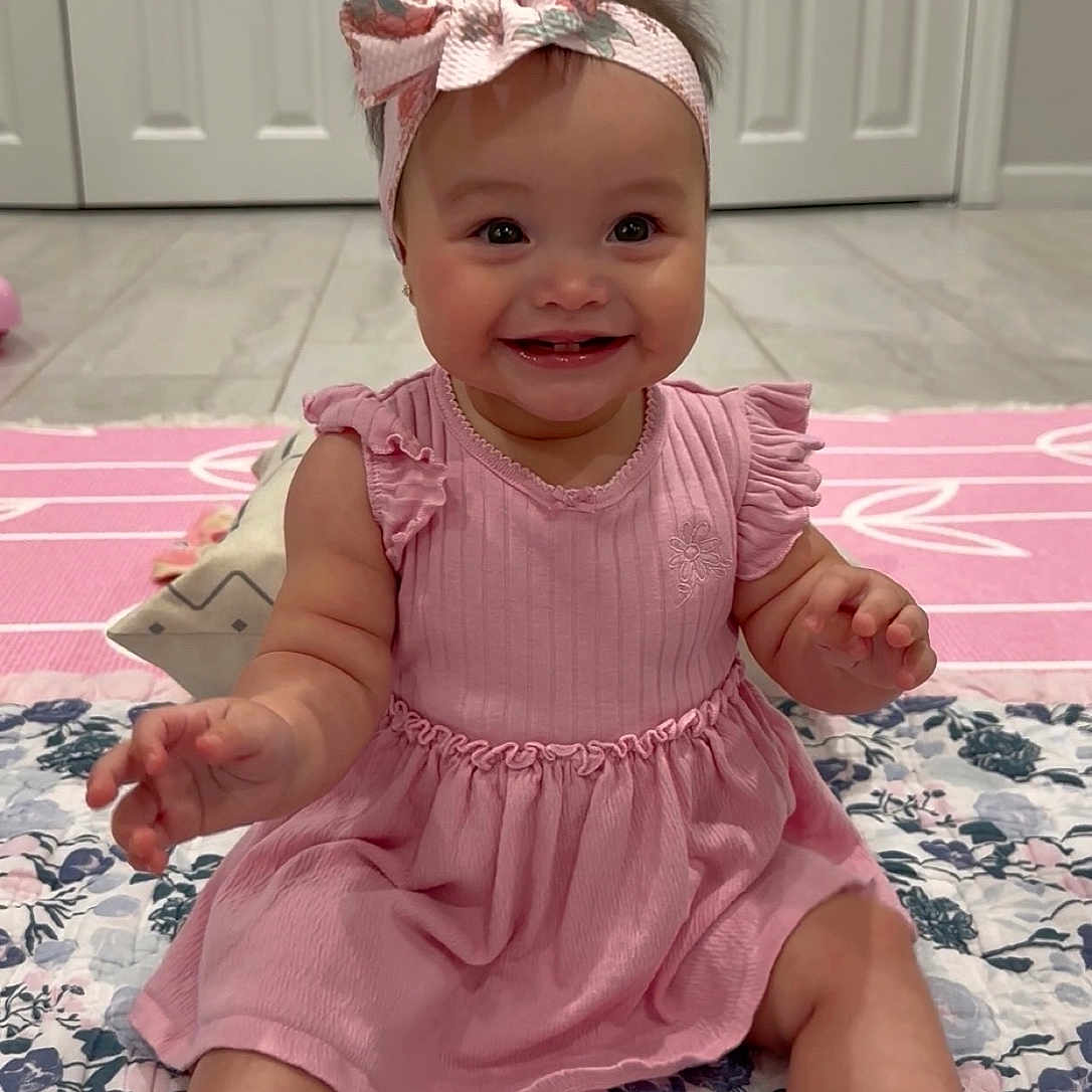 Eliana is registered to the contest to win money with this photo: arms, baby, bow, child, cute, eyes, face, floor, floral_pattern, girl, hair, happy, headband, indoors, mouth, pink_dress, quilt, sitting, smiling, toes