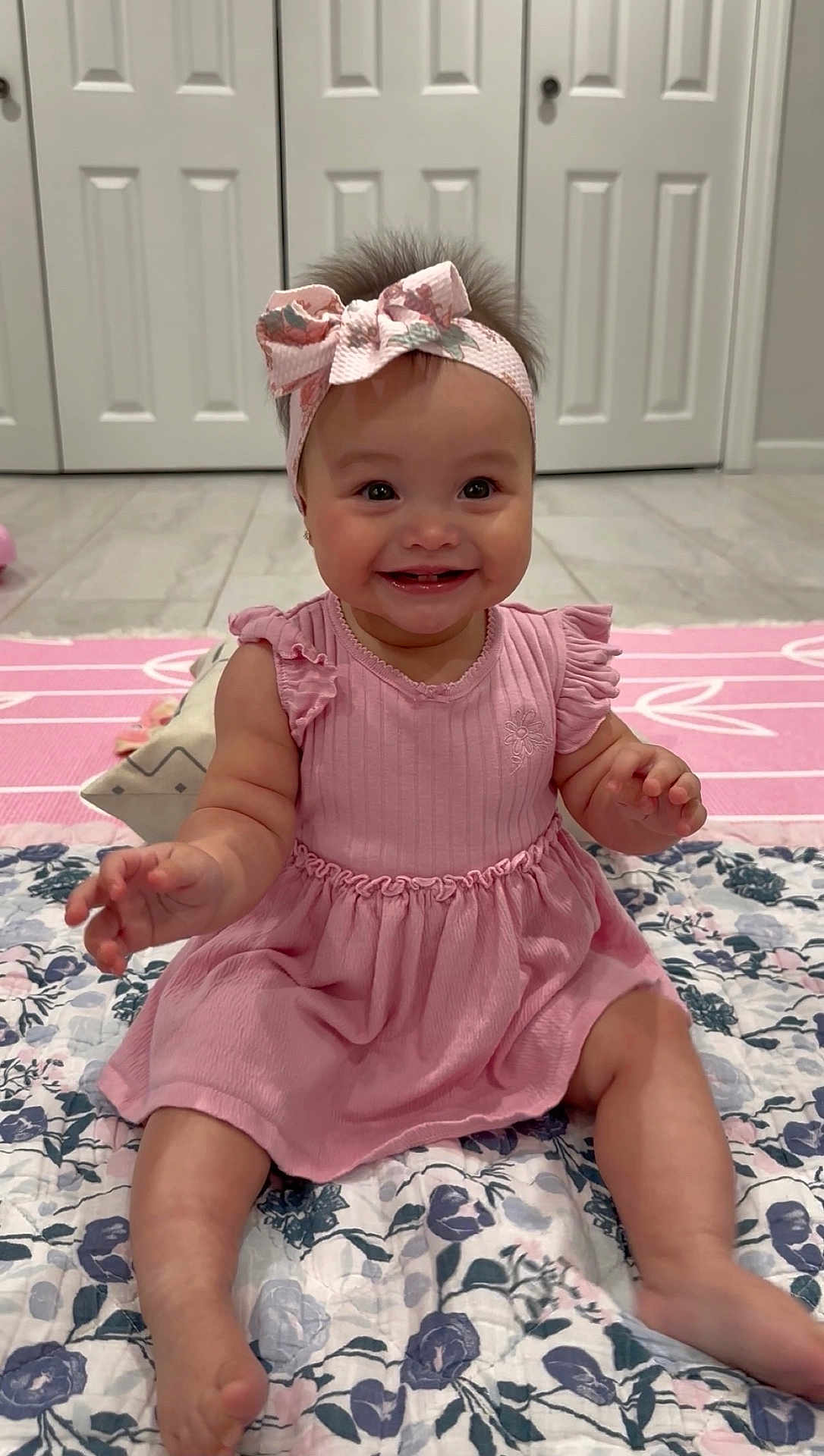 Eliana is registered to the contest to win money with this photo: baby, child, girl, smiling, pink_dress, headband, bow, sitting, floor, quilt, floral_pattern, indoors, happy, cute, toes, arms, face, eyes, hair, mouth