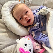 Neriah is registered to the contest to win money with this photo: baby, infant, pajamas, plush_toy, pink_bow, blanket, baby_seat, cushion, smiling, happy, face, person, indoor, soft_toy, cozy, child, cute, resting, comfort, wearing_mittens