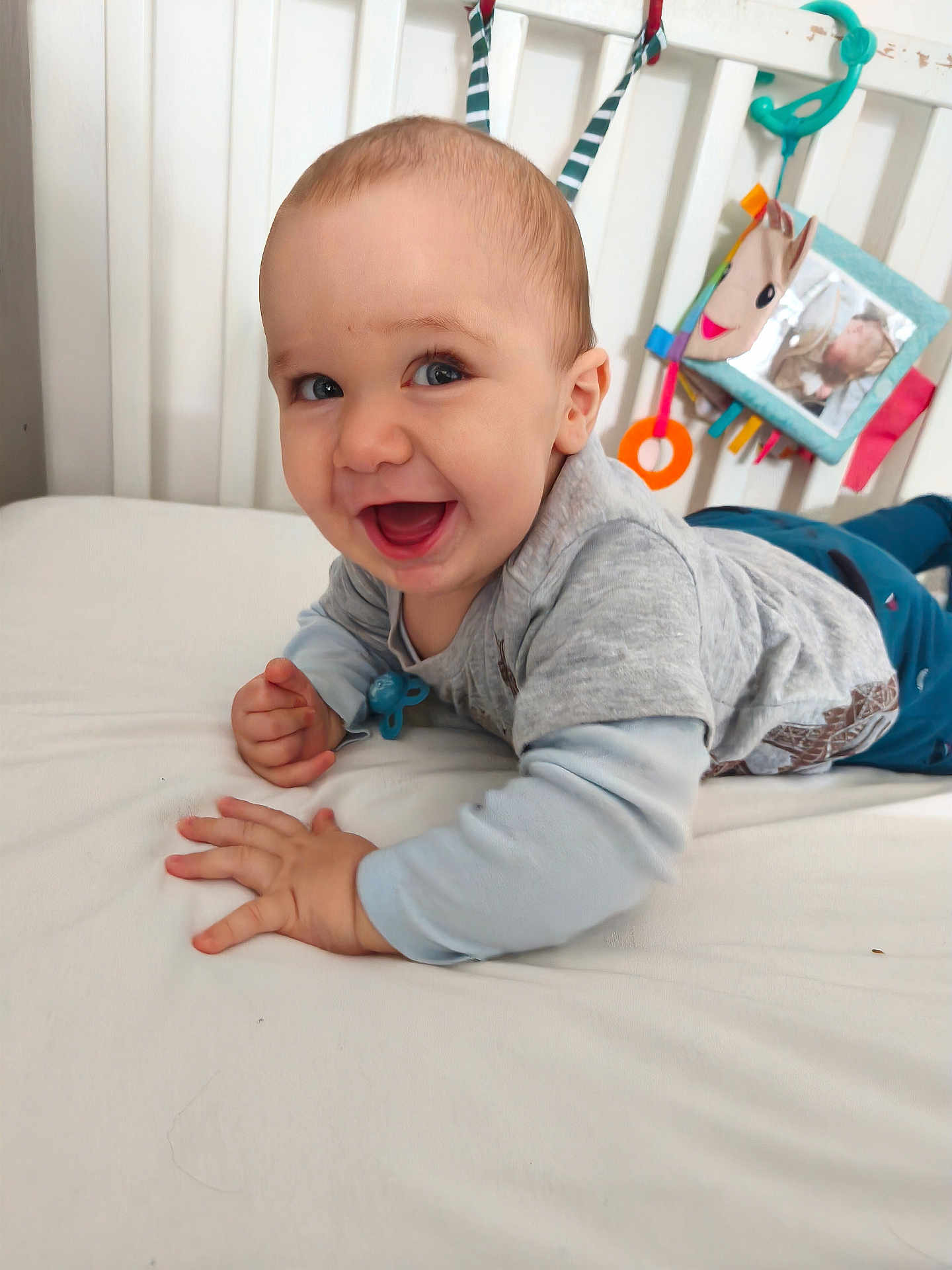 Milo participe au concours pour gagner de l'argent avec cette photo : baby, smiling, infant, bed, crib, toy, blue_clothing, gray_shirt, child, happy, indoor, nursery, cute, playful, young_child, pale_skin, light_hair, toddler, person, face