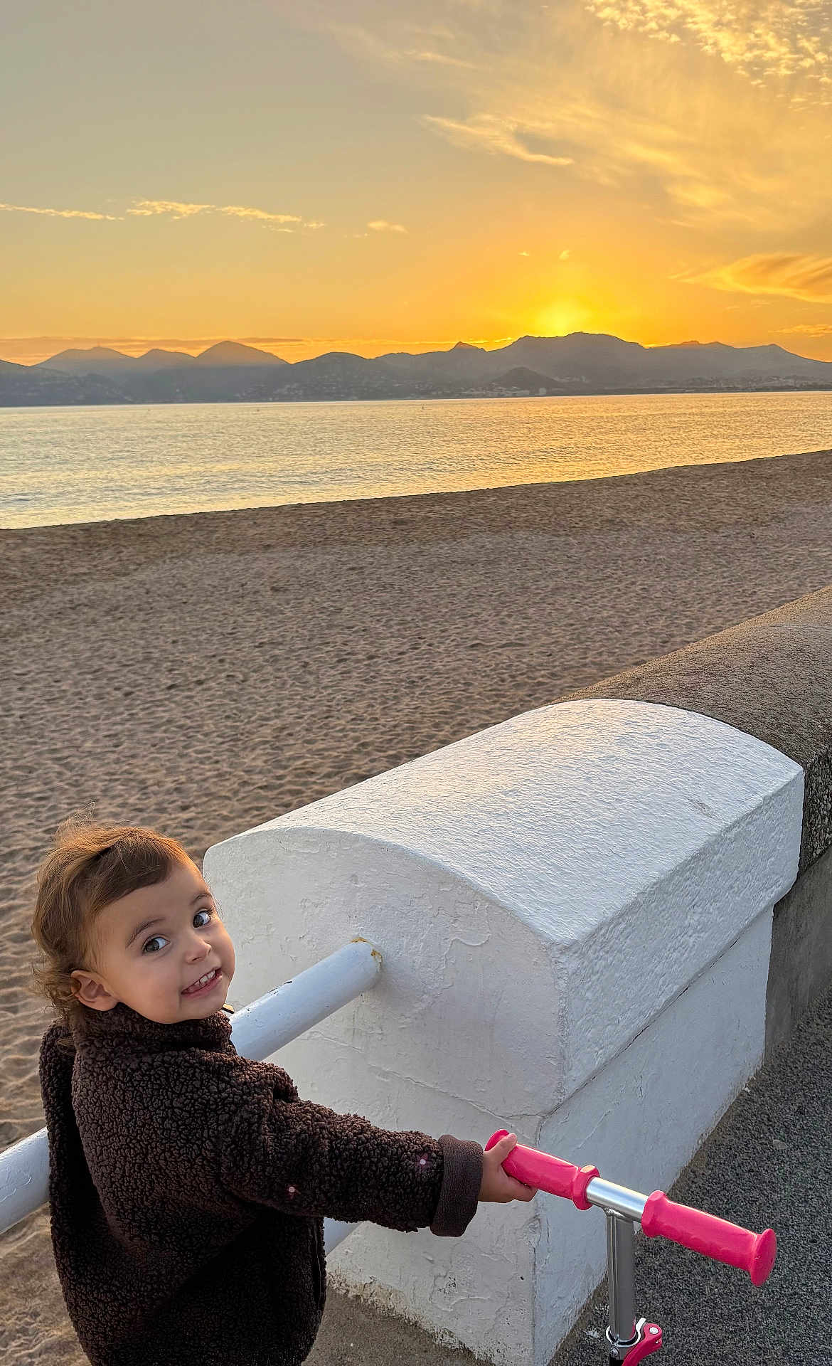 Carmela participe au concours pour gagner de l'argent avec cette photo : toddler, child, smile, jacket, scooter, pink, beach, sunset, mountains, water, sand, outdoor, person, happy, scenic, sunlight, nature, sky, play, leisure