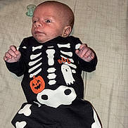 Wyatt is registered to the contest to win money with this photo: baby, infant, child, skeleton_costume, halloween, ghost, onesie, black_clothing, white_bones, pacifier, bed, blanket, lying_down, portrait, face, hands, feet, indoors, cozy, surprised