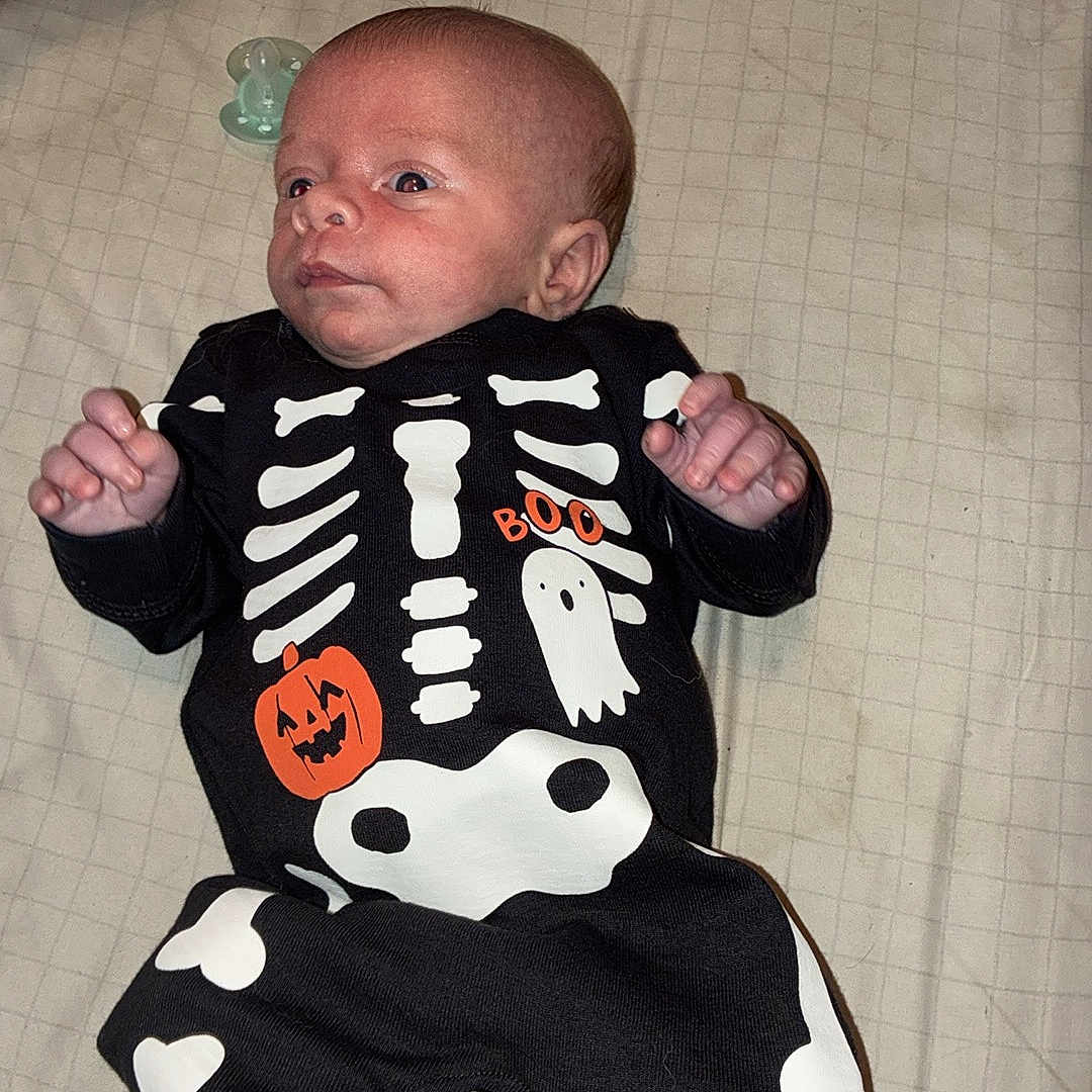 Wyatt is registered to the contest to win money with this photo: baby, infant, child, skeleton_costume, halloween, ghost, onesie, black_clothing, white_bones, pacifier, bed, blanket, lying_down, portrait, face, hands, feet, indoors, cozy, surprised