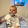 baby, pacifier, child, white_outfit, blue_floral_pants, sitting, indoor, face, hand, toy, comfort, television, blurred_background, cute, young_child, happy, portrait, soft_light, home, room