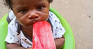 Zyaire is registered to the contest to win money with this photo: baby, casual_clothing, child, closeup, curly_hair, cute, food, green_seat, hand, happy, infant, outdoor, person, popsicle, red, sitting, skin, snack, summer, toddler