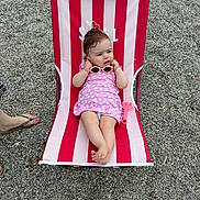 Mia a rejoint le concours — aidez-le/la à gagner de superbes lots ! baby, child, sunglasses, beach_chair, striped, pink_dress, outdoor, pebbles, footwear, sandal, person, summer, relaxing, toy, cute, infant, casual, expression, barefoot, vacation