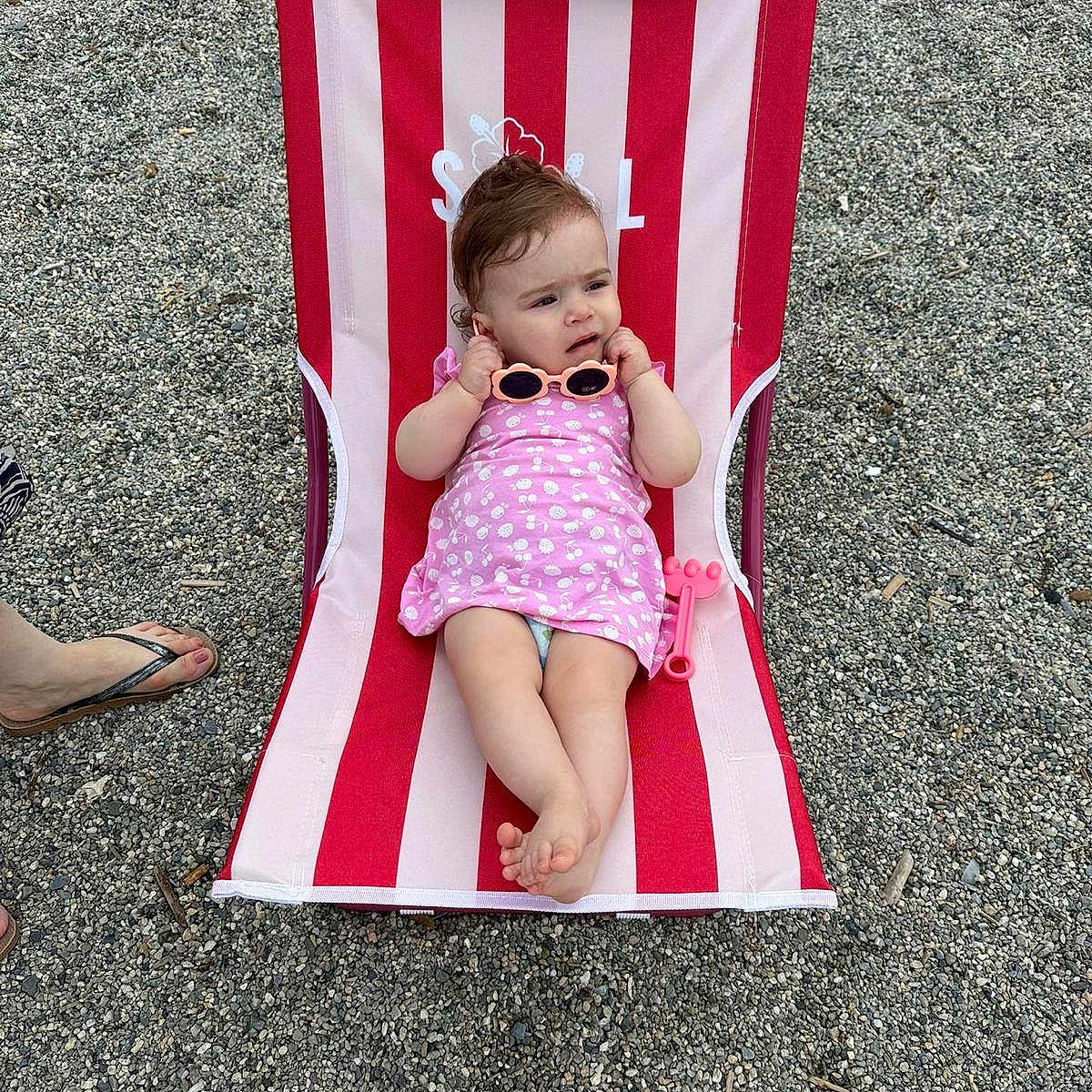 Mia a rejoint le concours — aidez-le/la à gagner de superbes lots ! baby, barefoot, beach_chair, casual, child, cute, expression, footwear, infant, outdoor, pebbles, person, pink_dress, relaxing, sandal, striped, summer, sunglasses, toy, vacation