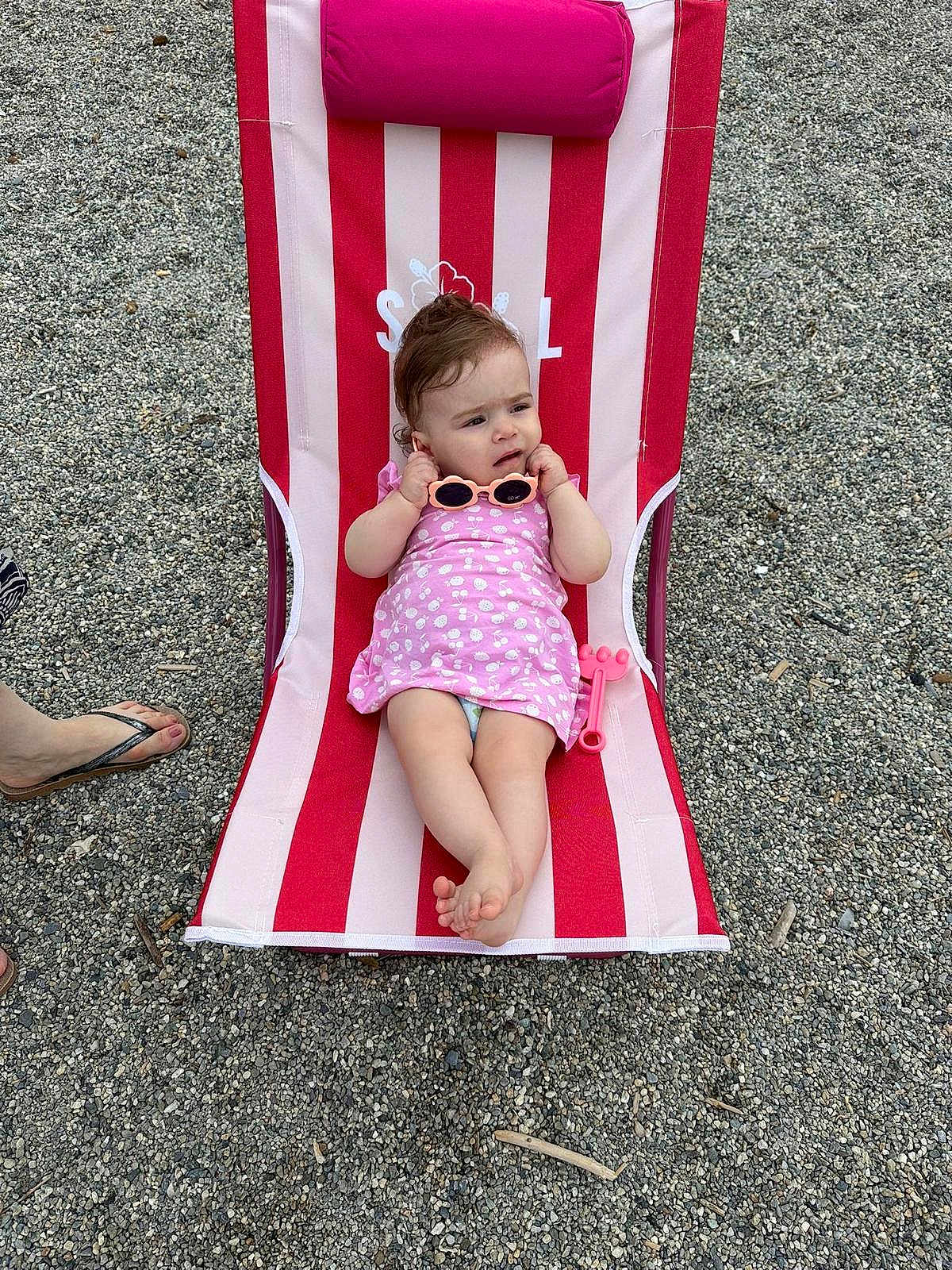 Mia a rejoint le concours — aidez-le/la à gagner de superbes lots ! baby, child, sunglasses, beach_chair, striped, pink_dress, outdoor, pebbles, footwear, sandal, person, summer, relaxing, toy, cute, infant, casual, expression, barefoot, vacation