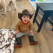 Archer is registered to the contest to win money with this photo: blanket, boots, brown_sweater, child, cowboy_hat, cute, dog, gray_pants, happy, home, indoor, living_room, person, playtime, sitting, smiling, table, toddler, toy, wooden_floor