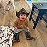 toddler, child, cowboy_hat, boots, brown_sweater, gray_pants, smiling, sitting, wooden_floor, dog, toy, table, blanket, indoor, happy, person, living_room, playtime, cute, home