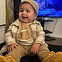 baby, infant, child, smile, beanie, earring, plaid_shirt, jacket, sneakers, shoes, television, subtitles, living_room, curtain, window_blinds, mom_sign, sitting, portrait, cozy, happy