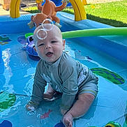 James is registered to the contest to win money with this photo: baby, child, swimsuit, inflatable_pool, water, bubbles, toy, green, yellow, blue, outdoor, grass, sunlight, play, fun, summer, animal_toy, smiling, sitting, backyard