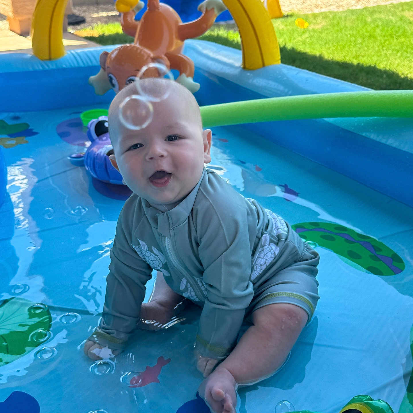 James is registered to the contest to win money with this photo: baby, child, swimsuit, inflatable_pool, water, bubbles, toy, green, yellow, blue, outdoor, grass, sunlight, play, fun, summer, animal_toy, smiling, sitting, backyard