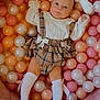 baby, baby_girl, ball_pit, child, colorful, cute_clothing, indoor, infant, orange_balls, pastel_balls, pink_balls, plaid_dress, play_area, portrait, relaxed, smiling_face, soft_toys, stuffed_animal, white_balls, white_socks