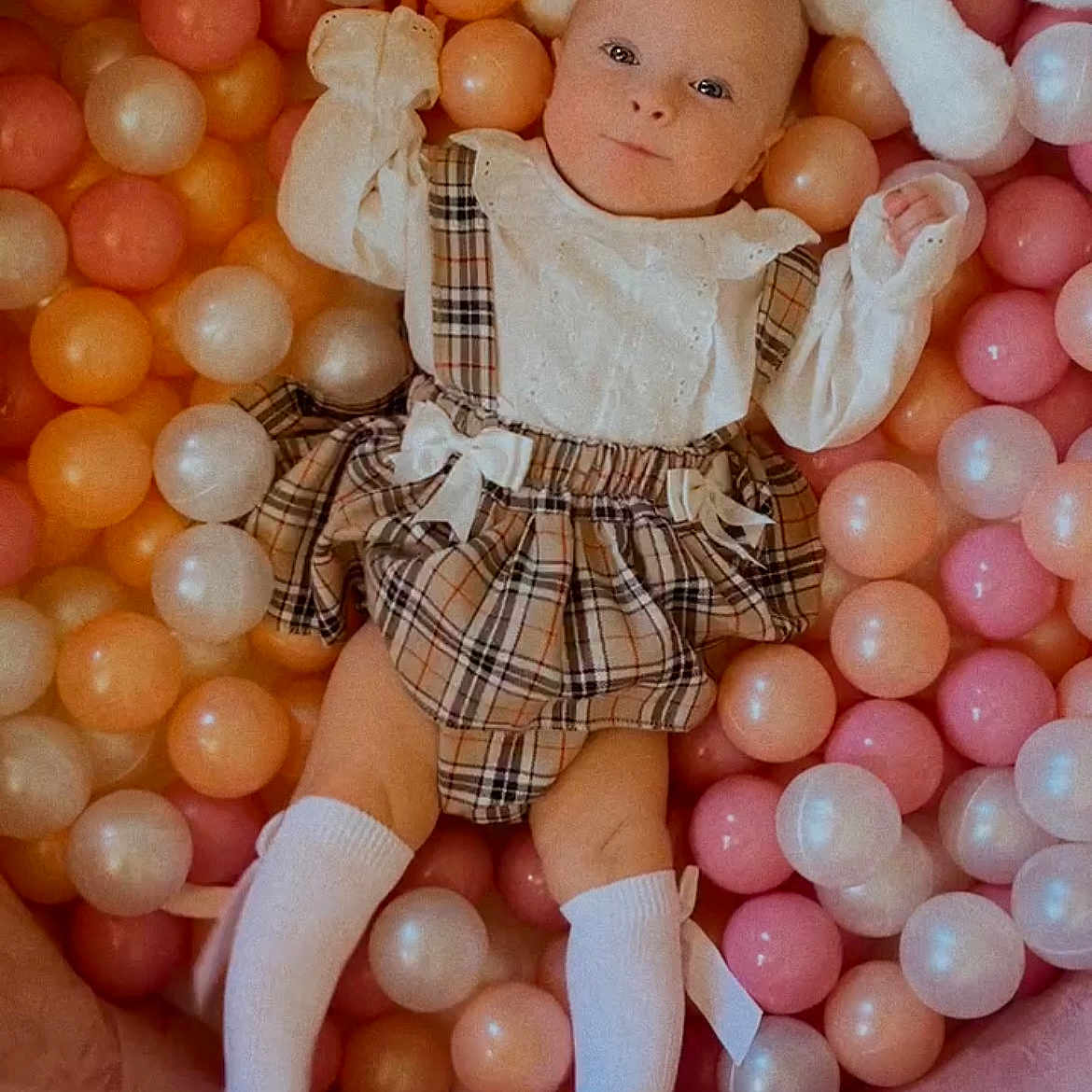 Phoebe-Rose is registered to the contest to win money with this photo: baby, baby_girl, ball_pit, child, colorful, cute_clothing, indoor, infant, orange_balls, pastel_balls, pink_balls, plaid_dress, play_area, portrait, relaxed, smiling_face, soft_toys, stuffed_animal, white_balls, white_socks