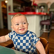 Theodore is registered to the contest to win money with this photo: child, toddler, smile, face, indoor, checkered_shirt, blue_shirt, short_hair, seated, table, wooden_chair, happy, curious, portrait, person, baby, cute, young_child, home, blurred_background