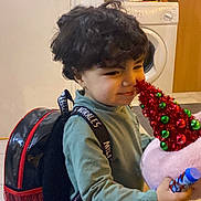 Ilyan participe au concours pour gagner de l'argent avec cette photo : child, curly_hair, backpack, plush_toy, party_hat, indoor, smile, green_clothing, table, washing_machine, mirror, floor_tiles, cabinet, toy, happy, person, home, curious, young_child, casual