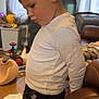 child, boy, white_shirt, black_pants, indoor, living_room, toys, remote_control, curtains, couch, wooden_floor, side_profile, leaning, young_child, casual_clothing, furniture, home, contemplative, person, footwear