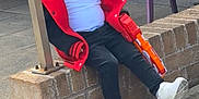 Jaxon is registered to the contest to win money with this photo: child, sitting, red_jacket, white_shirt, black_pants, white_sneakers, toy_gun, brick_wall, outdoor, pavement, purple_chairs, person_in_jeans, playful, casual, daylight, young, happy, relaxed, concrete, urban