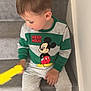 child, boy, stairs, mickey_mouse, sweatshirt, gray_pants, white_shoes, carpet, indoor, toy, sitting, looking_down, casual_clothing, young, side_view, hand, footwear, wall, home, portrait