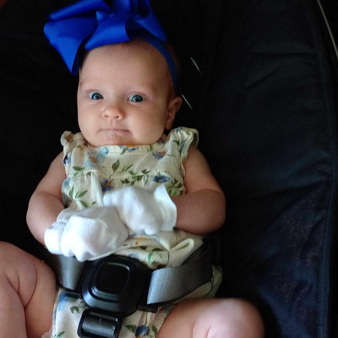 Peyton is registered to the contest to win money with this photo: arms, baby, baby_seat, black_seat, blue_bow, child, closeup, comfortable, curious_expression, cute, drool, floral_outfit, headband, indoor, infant, legs, mittens, person, sitting, straps