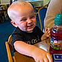 toddler, child, toy_train, table, indoor, smiling, blonde_hair, black_shirt, wooden_furniture, blue_chair, person, happy, play, dining_area, casual, background_blur, face, arm, hand, juice_bottle